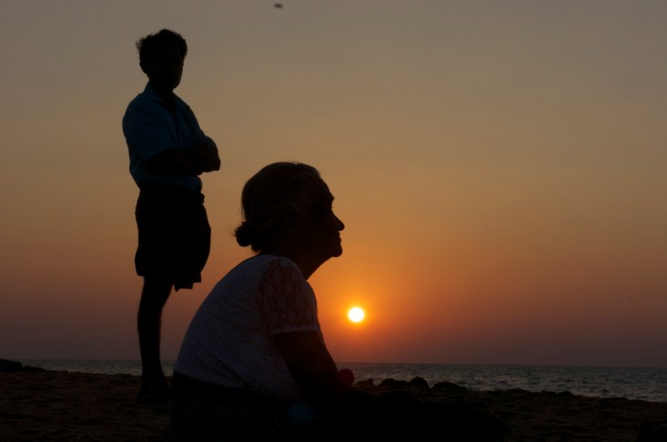 een oude vrouw geniet met haar zon op het strand van de ondergaande zon, een passend beeld bij het werk van Medi-Aid. 